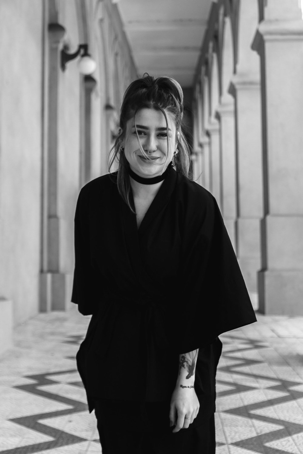 retrato em preto e branco da nat, fundadora da dóiner, de roupa preta e gargantilha, sorrindo num corredor de arcos